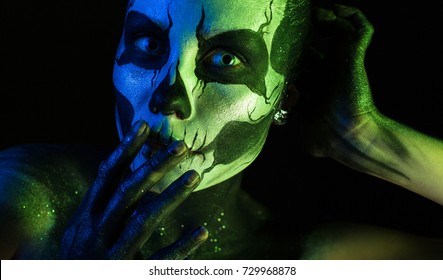 Isolated on black, closeup picture, toned green and blue, beautiful 
creepy young blonde caucasian woman with scull body art, grey eyes, hands on head, look at camera