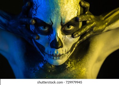 Isolated on black, closeup picture, toned yellow and blue, spooky young blonde caucasian pretty woman with scull body art, grey eyes, stretches her face with her hands, look at camera