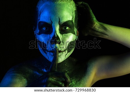 Isolated on black, closeup picture, toned green and blue, beautiful 
creepy young blonde caucasian woman with scull body art, grey eyes, hand on head, points to be silent