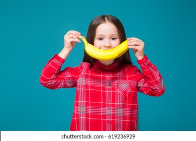 Isolated On Blue, Attractive Caucasian Brunette Child In Red Plaid Shirt And Blue Jeans With Long Hair Hold Bananas Near Her Face, Look At Camera