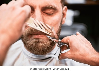 Isolated photo of professional hairdresser using comb and scissors to shape mustache. Male customer on appointment in a barbershop. Barber shaving thick beard. - Powered by Shutterstock