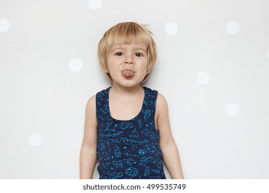 Isolated portrait of adorable funny baby boy with blue eyes wearing tank top having fun indoors, showing his tongue at camera, teasing, standing against studio wall with copy space for your content