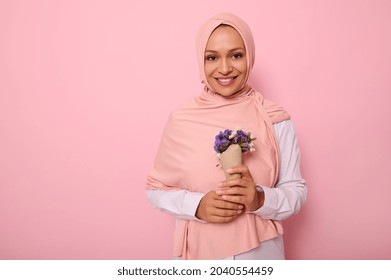 Isolated portrait on colored background of a beautiful Muslim Arab woman in pink hijab, holding a bouquet of wildflowers in purple shades, wrapped in craft paper, smiles at camera with a toothy smile
