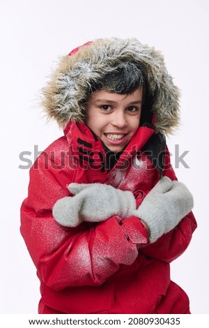 Isolated portrait on white background of a handsome Caucasian elementary aged boy, dressed in a bright red parka a snow-covered hood, looking at the camera while hugging himself shivering from cold