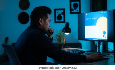 Isolated Portrait Of A Young Asian Animator And Graphic Design Focused On Developing A Website. Wearing A Fancy Blue Turtleneck, Hand On His Chin Thinking, Background Is A Dark Bedroom With Paintings.