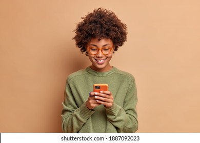 Isolated shot of African American woman uses smartphone application enjoys browsing social media creats news content makes online order wears spectacles and casual jumper poses over beige studio wall - Shutterstock ID 1887092023