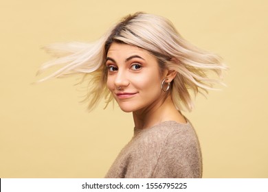 Isolated shot of beautiful amazing young female in stylish oversize sweater turning around with blonde hair flowing in wind, smiling at camera, advertising volumising shampoo. Beauty and hair care