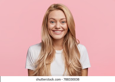 Isolated shot of beautiful blonde female model with even teeth and shining smile, has bugged blue eyes, has good festive mood, being in anticipation of coming winter holidays, isolated on pink