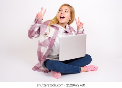 Isolated shot of cheerful Beautiful caucasian teen girl sitting with laptop in lotus position on white background makes peace or victory sign with both hands, feels cool.