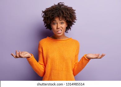 Isolated shot of confused beautiful woman with dark skin, curly haircut, spreads hands sideways, smirks face, feels doubt while makes choice, dressed in casual jumper, isolated over purple wall