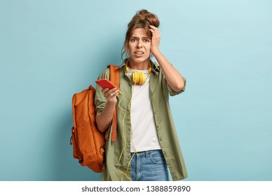 Isolated Shot Of Dissatisfied Schoolgirl Holds Cell Phone Connected To Headphones, Has Frustrated Expression, Can Not Listen Music Without Internet. Oh No, My Songs Disappeared From Playlist