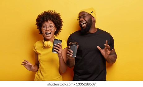 Isolated shot of energized overjoyed dark skinned teenagers dance actively in rhythm of music, have spare time for drinking coffee, wear casual t shirts, spectacles, isolated over yellow studio wall