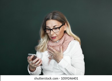 Isolated Shot Of Fashionable Senior Woman With Blonde Hair Holding Hand On Her Chest And Opening Mouth In Shock, Stunned With Bad Negative News That She Reads Via Text Message On Smart Phone
