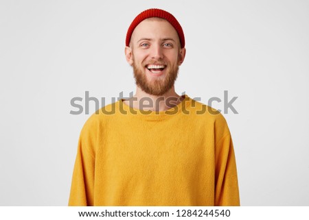 Isolated shot of joyful bearded young cute man laughs joyfully as hears funny joke, wears red hat and sweater, poses against white studio wall. Happiness and positive emotions concept