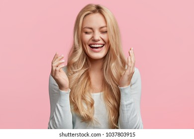 Isolated shot of joyful blonde young cute woman laughs joyfully as hears funny anecdote from friend, has long light hair, poses against pink studio wall. Happiness and positive emotions concept