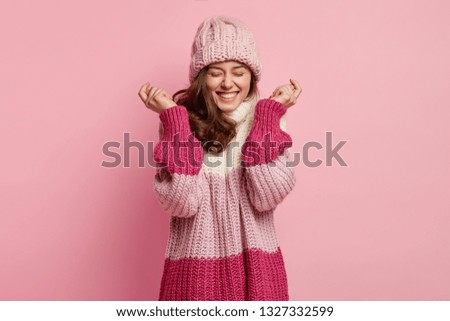 Isolated shot of overjoyed woman raises hands, wears knitted hat and loose sweater, expresses positive emotions, laughs sincerely at funny story, isolated over pink background. Emotions concept