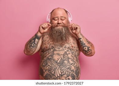 Isolated shot of overweight bearded man curls mustache, closes eyes, listens favorite songs in headphones, found music station or funny podcast, has tattooed naked belly, models against rosy wall