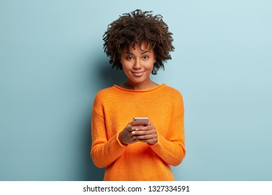 Isolated shot of positive smiling woman holds modern cellular, enjoys wireless internet, checks bank account or sends message in social networks, wears orange clothes, models over blue background - Powered by Shutterstock