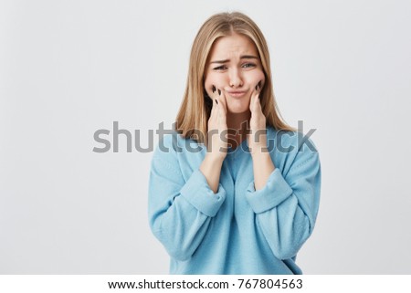 Isolated shot of pretty cute blonde girl in blue clothes touching her cheeks with her hands, showing how big they are. Funny female mocking and posing against studio wall.