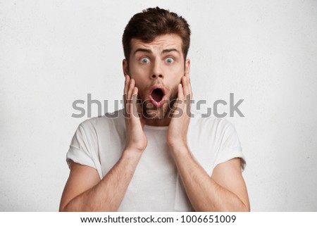 Isolated shot of surprised blue eyed male has beard, dressed in casual white t shirt, looks with bugged eyes, being shocked to be invited on business meeting with partners, doesn`t know what to wear