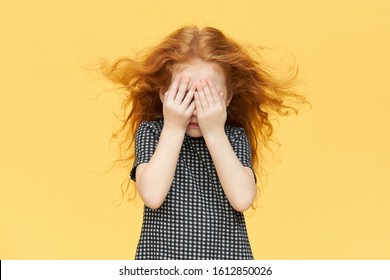 Isolated Shot Of Timid Little Girl Being Shy, Covering Eyes. Red Haired Female Child Hiding Tears Behind Her Hands, Crying. Caucasian Kid Playing Seek And Hide Against Blank Yellow Wall Background