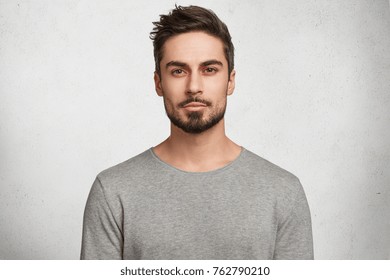 Isolated shot young handsome male and beard  mustache   trendy hairdo  wears casual grey sweater  has serious expression as listens to interlocutor  poses in studio against white background