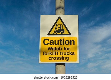 Isolated Sign Caution Watch For Fork Lift Trucks Crossing With Yellow Warning Triangle And Icon Of Man In Fork Lift Truck. Sunny Day With Blue Sky And Light Cloud. UK