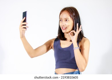 Isolated Studio Cutout Shot Of Asian Young Female Teenager Model In Crop Top Shirt And Jeans Smiling Greeting Hold Smartphone Video Call Online Via Internet Showing Okay Hand Sign On White Background.