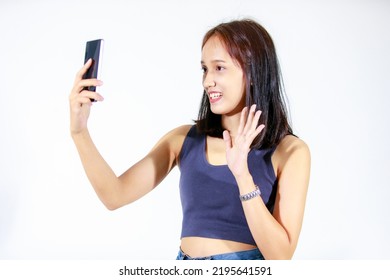 Isolated Studio Cutout Shot Of Asian Young Female Teenager Model In Crop Top Shirt And Jeans Smiling Greeting Hold Smartphone Video Call Online Via Internet Showing Okay Hand Sign On White Background.