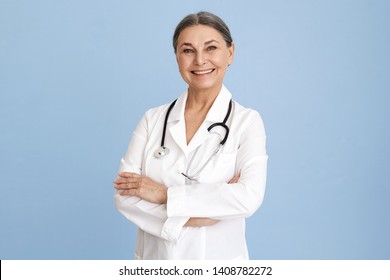 Isolated studio image of charismatic beautiful Caucasian senior female chief doctor crossing arms on her chest confidently and smiling broadly dressed in white overall with stethoscope around her neck