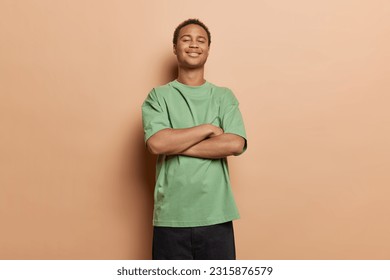 Isolated studio shot of dark skinned young man stands self confident keeps arms folded dressed in casual green t shirt and black trousers feels proud of himself poses against brown background.