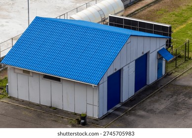 Isolated view of compact utility building featuring vivid blue metal roof and matching doors. Clean geometric shape and industrial surroundings emphasize functional architecture and service-facility a