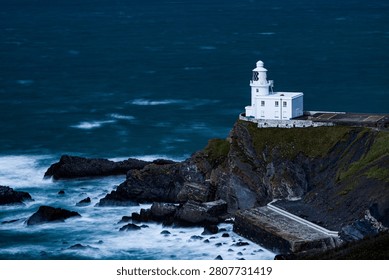 Isolated white lighthouse on a dramatic rocky headland at dusk, deep blue sea with white surf breaking against black rocks below, moody twilight tone, North Devon coast.