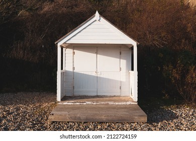 An Isolated Wooden White Beach Hut With A Porch And Decking On A Pebble Beach In Warm Evening Light At Cooden In East Sussex, United Kingdom