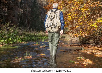 Isolated From Work, Seniors Finally Have Time. Hiking Is A Popular Pastime For Many Seniors. Older Man With Waders Wanders Through The Still Small River Neckar In The Neckar Valley.