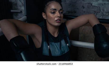 Isolated Young Ethnic Female Athlete Sitting At The Corner of a Boxing Ring Resting In Between Sparring Sessions and Training With Her Coach For The Championships. Strong Confident Female Kick Boxer. - Powered by Shutterstock