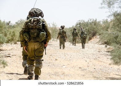 Israeli Army Combat Soldiers Return To Base After Completing Military Mission During Military Campaign / War. Desert Landscape, Armed Infantry Soldier Walking In The Foreground. Soldiers Walking Away.