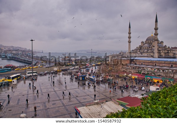 Istanbul, Turkey - December 31, 2013: View from\
above of the crowd of people, boats and cars with  Galata bridge,\
New Mosque and Bosporus strait in the back, in Istanbul, Turkey on\
a cloudy day.