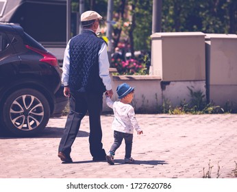 Istanbul, Turkey - May 7 2020: Cute Little Toddler Boy With A Stylish Hat Is Holding The Hands Of His Grandfather While Walking. Grandpa Wears Masks And Grandson Enjoys The Walk Next To Him.