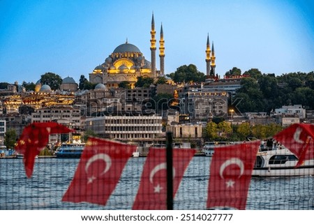 Türkiye. Istanbul. View of the Süleymaniye Mosque, illuminated at dusk, with Turkish flags flying in the foreground and boats sailing on the Bosphorus.