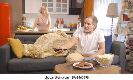 Istanbul,Turkey-01.27.2021:While the old woman is preparing the table in the kitchen, her husband is lying on the sofa, watching important things on the TV and eating popcorn. 