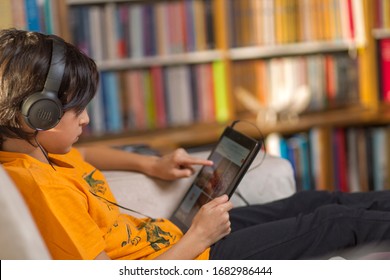 Istanbul-Turkey/March 23 2020: Teenage Dressed In Orange T-shirt Is Using Digital Tablet With Black Headphones. Bookcase In Background. Stay At Home. Youth, Technology And Online Concepts. Horizontal.