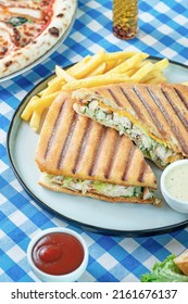 An italan sandwich called panini that contains chicken, lettuce in it, frenche fries and tiny bowls of ketchup and garlic sauce into a white plate on a white and blue plaid tablecloth.