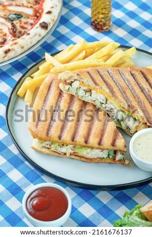 An italan sandwich called panini that contains chicken, lettuce in it, frenche fries and tiny bowls of ketchup and garlic sauce into a white plate on a white and blue plaid tablecloth.