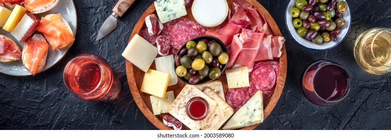 Italian antipasti or Spanish tapas panorama. Gourmet cold meat and cheese platter on a table, shot from the top with wine on a black background. A variety of appetizers