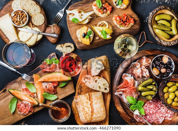 Italian antipasti wine snacks set. Brushettas,\
cheese variety, Mediterranean olives, pickles, Prosciutto di Parma\
with melon, salami and wine in glasses over black grunge\
background, top view