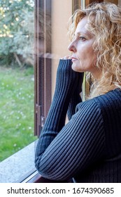 Italian Blonde Woman Forced Into House By Quarantine Due To Coronavirus Covid-19, Looks At The Outside World From The Window Of Her Home With Bored Expression