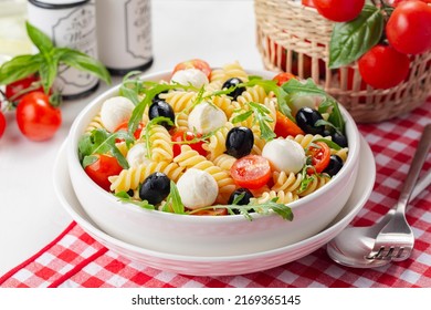 Italian cold pasta salad or Pasta fredda alla caprese. Fusilli, tomato, mozzarella, olive, arugula. White bowl, fresh vegetables on background.