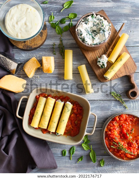 Italian cuisine. Cannelloni with spinach and\
ricotta. Cannelloni is prepared with parmesan cheese in tomato\
sauce. Italian dinner. Italian food on a gray wooden background.\
Bechamel sauce