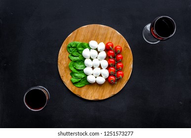 Italian cuisine, food concept. Italian flag made of mozzarella, tomatoes, basil on wooden cutting board near glass of red wine on black background top view copy space